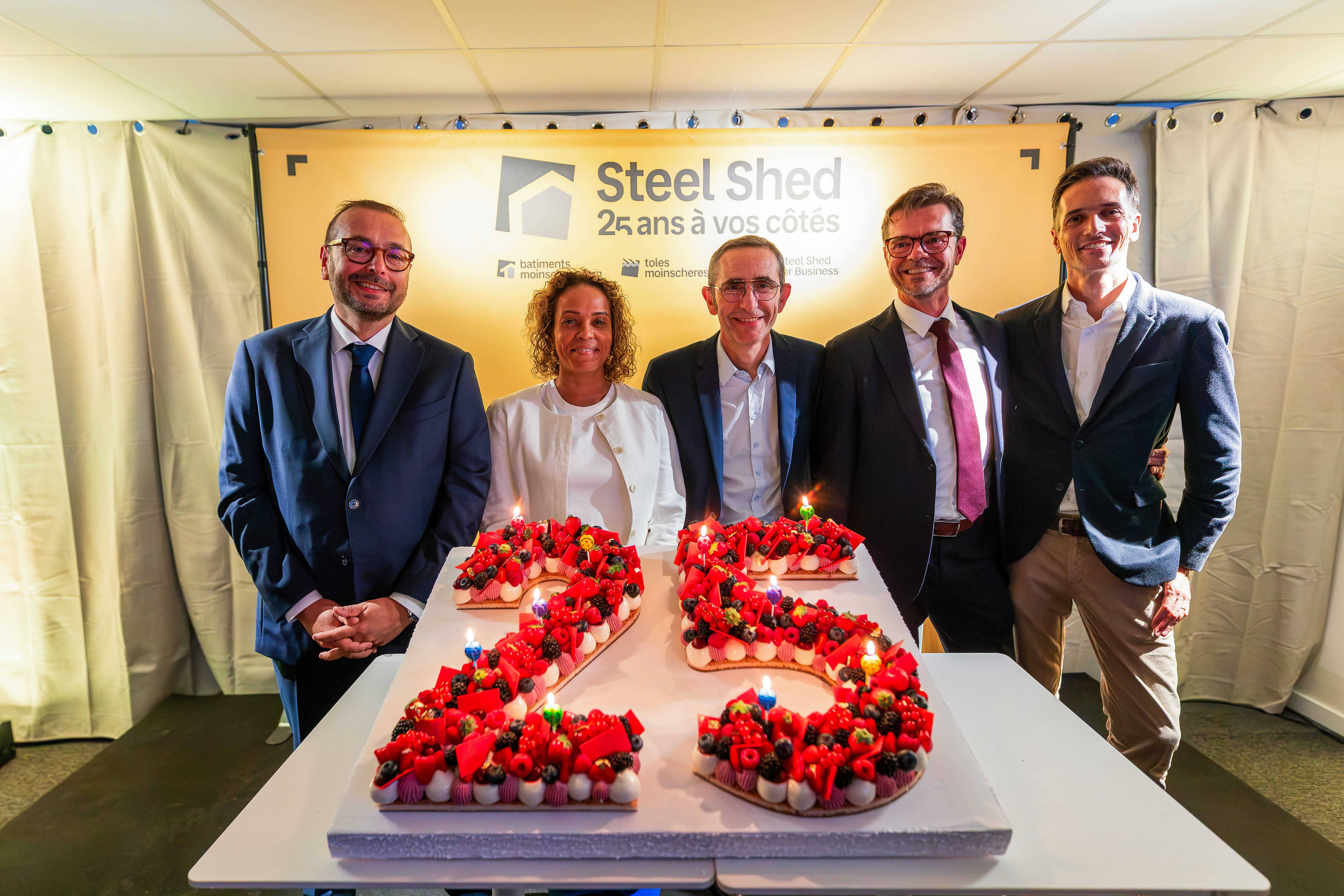 Cinq personnes posent en souriant derrière un gâteau en forme de 25 décoré de fruits rouges, devant une bannière jaune « Steel Shed, 25 ans à vos côtés » portant les logos batimentsmoinschers.com, tolesmoinscheres.com et Steel Shed for Business.